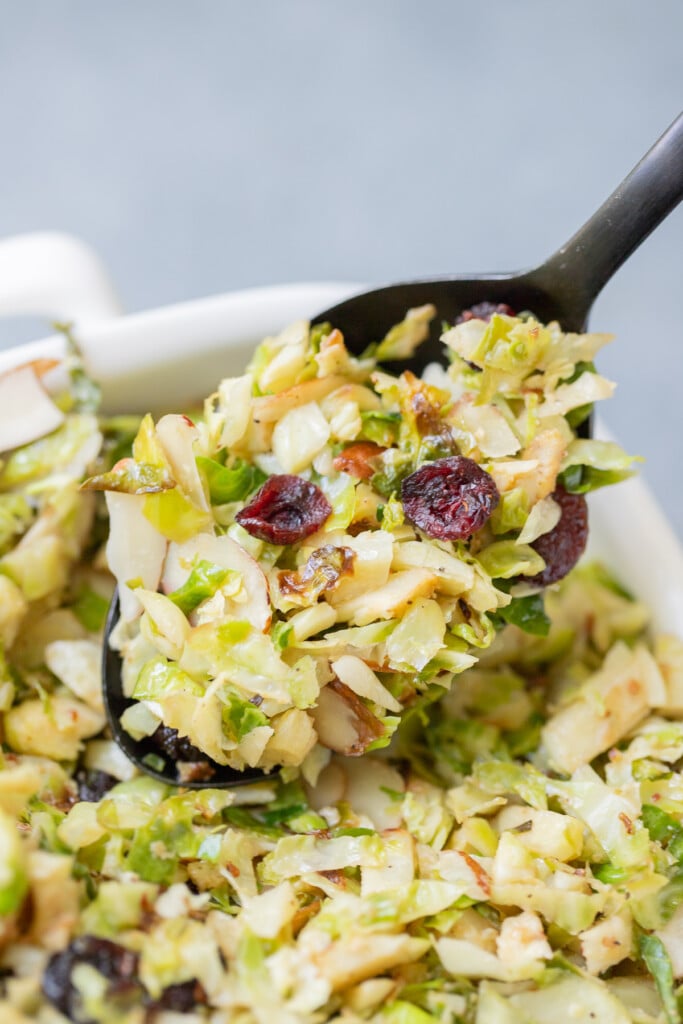 Shaved Brussels Sprouts Salad in a white dish on a grey background. A black spoon is holding up a scoop of the salad.