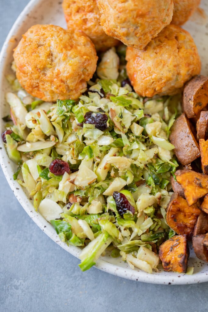 shaved brussels sprouts salad on a white plate with meatballs and roasted sweet potatoes on the side on a grey background
