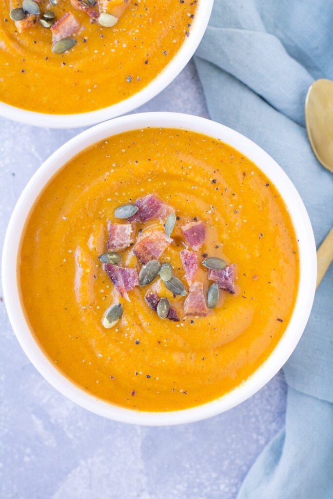 An overhead shot whole30 butternut squash soup in a white bowl on a grey background with a blue linen