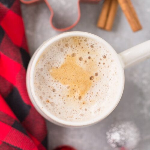 An overhead shot of a healthy gingerbread latte on a grey background with a red checked linen, ornaments and cinnamon sticks off to the side.