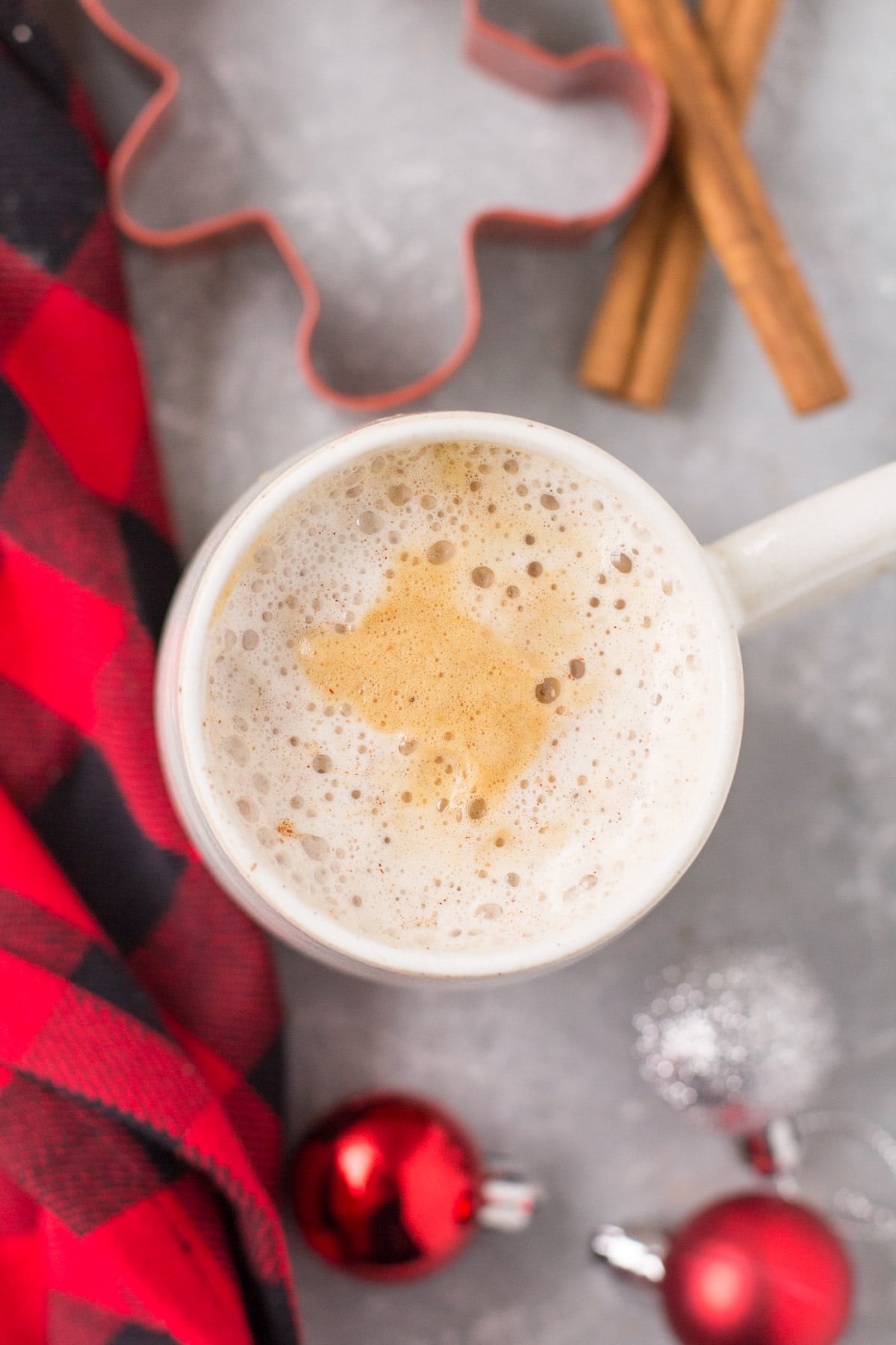 An overhead shot of a healthy gingerbread latte on a grey background with a red checked linen, ornaments and cinnamon sticks off to the side.