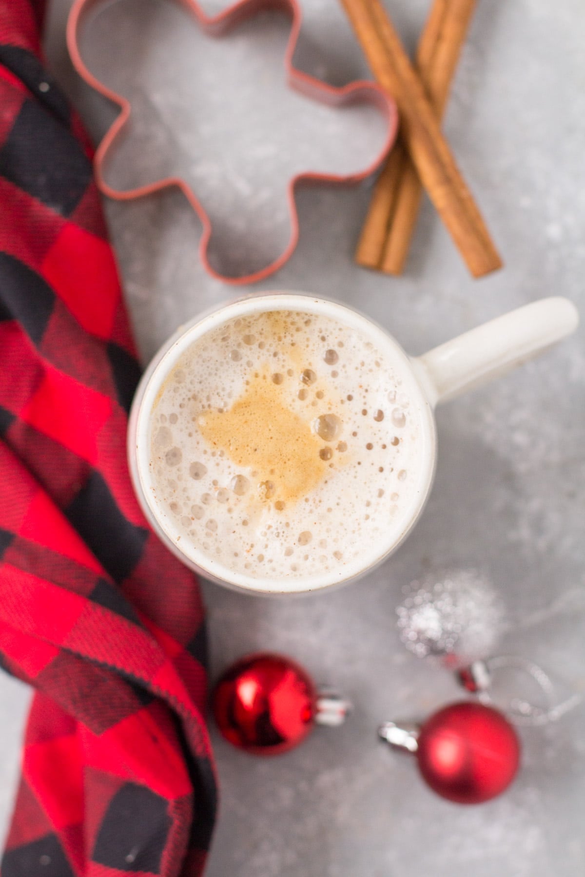 An overhead image of a healthy gingerbread latte with a red checked linen, ornaments, gingerbread cookie cutter + cinnamon sticks off to the side.