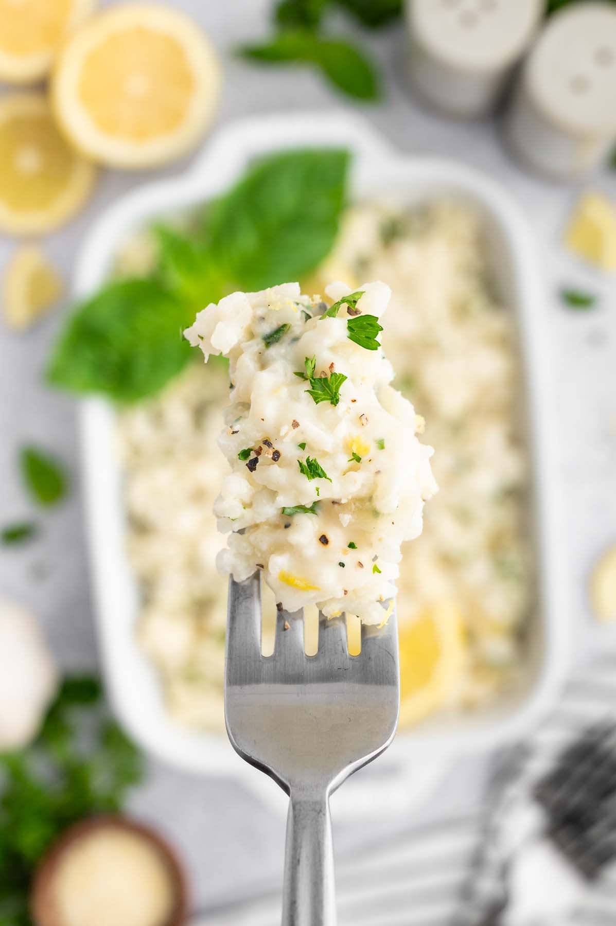 A fork holding up a bite of lemon risotto with a white dish of the risotto in the background.