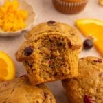 Healthy Cranberry Orange Muffins stacked up with a muffin with a bite out of it on the top with cranberries and oranges in the background.