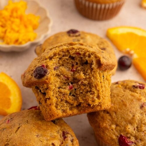 Healthy Cranberry Orange Muffins stacked up with a muffin with a bite out of it on the top with cranberries and oranges in the background.