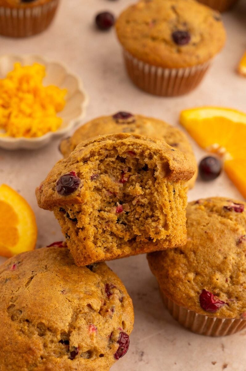 Healthy Cranberry Orange Muffins stacked up with a muffin with a bite out of it on the top with cranberries and oranges in the background.