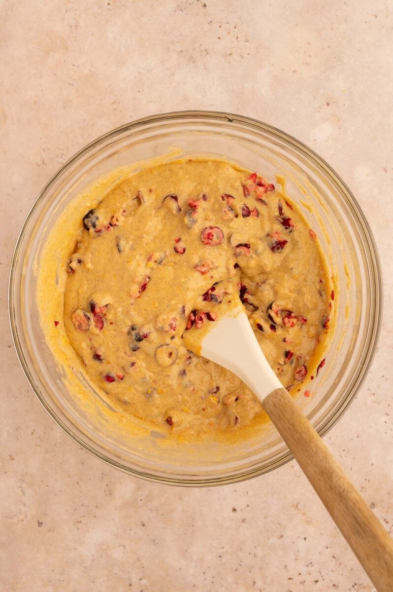 Cranberries folded into an orange muffin batter in a clear bowl with a rubber spatula in it.