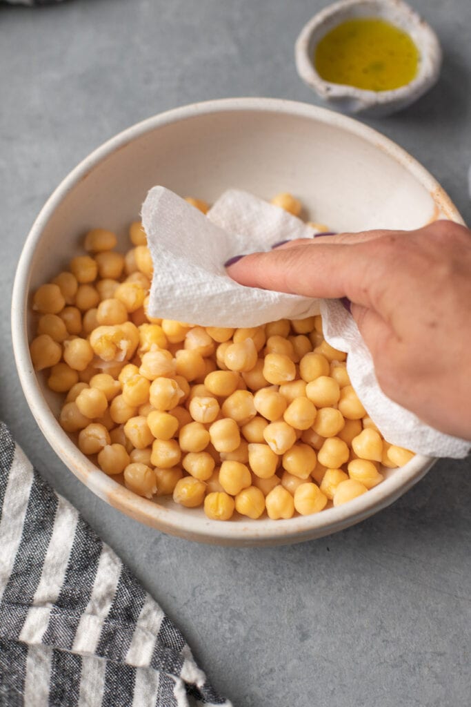 a hand patting chickpeas in a bowl dry with a paper towel