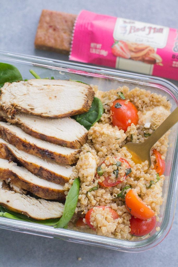 A close up image of Chicken Caprese Quinoa Meal Prep Bowls with a gold fork taking a bite out of it + a granola bar off to the side