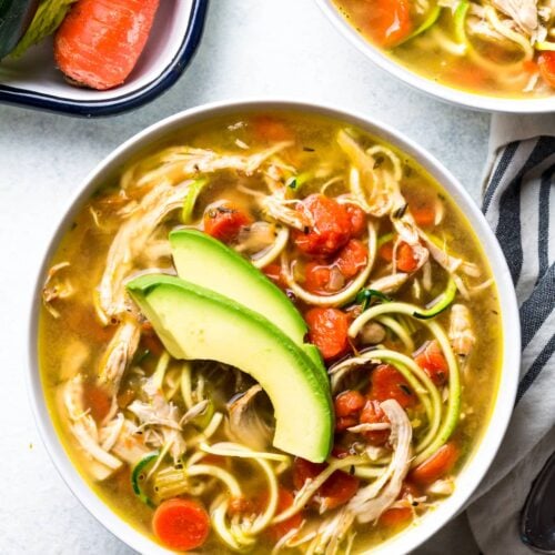A bowl of Paleo Chicken Soup on a grey background with veggies and a linen off to the side
