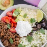 A close up image of taco cauliflower rice bowls in a white bowl with an avocado and yogurt off to the side.