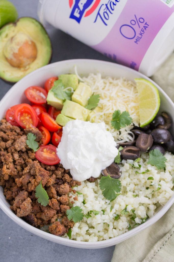 A close up image of taco cauliflower rice bowls in a white bowl with an avocado and yogurt off to the side.