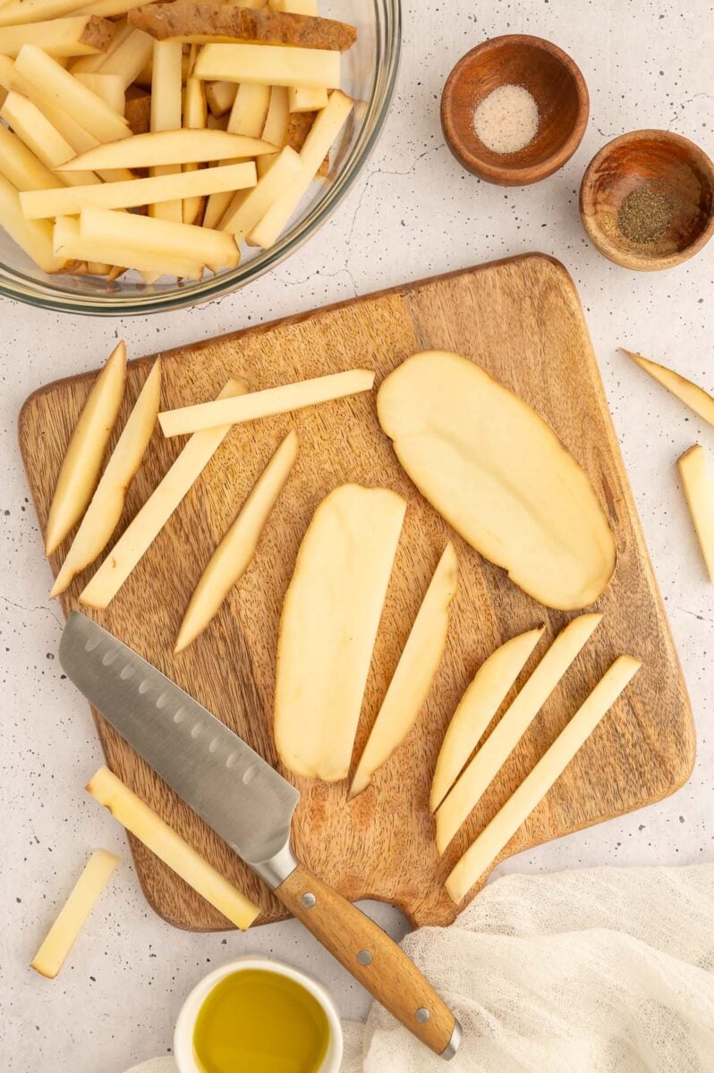 A cutting board with potatoes cut into slabs and then into sticks for baked french fries and a bowl of the fries on the side.