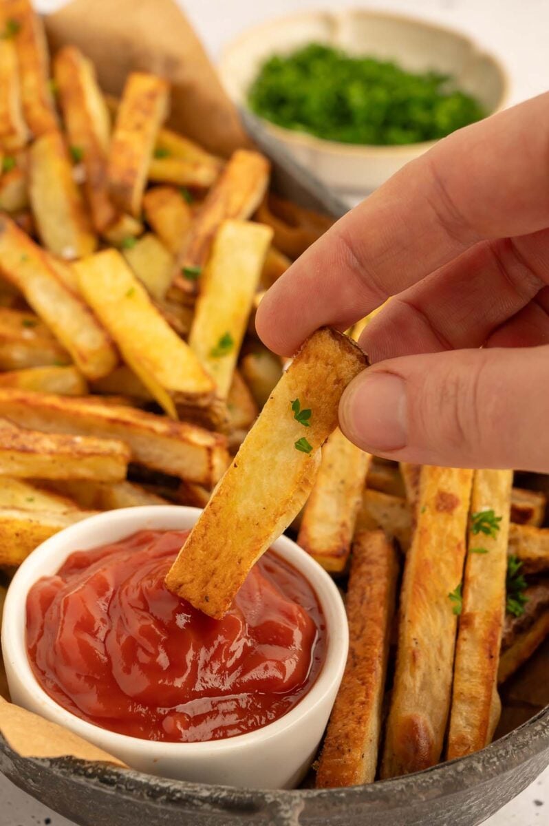 A single baked french fry being dipped in a small bowl of ketchup with a platter in the background.