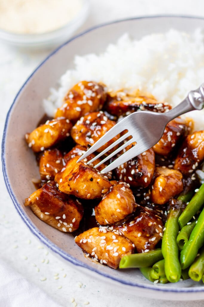 a plate of healthy sesame chicken, white rice and green beans with a gold fork taking a bite out of it.