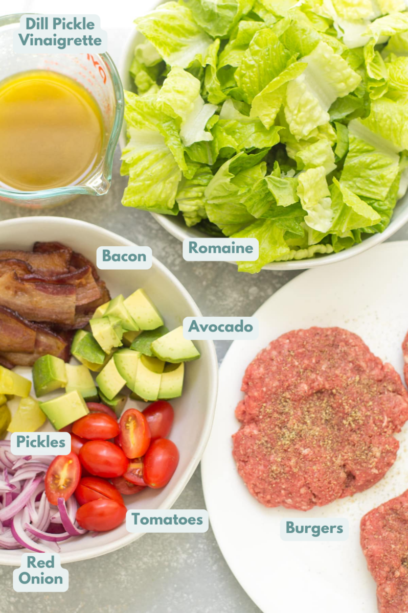 Ingredients for a burger salad laid out in bowls on a grey background: ground beef patties, crispy bacon, cherry tomatoes, romaine lettuce, onions, pickles, avocado, dill pickle salad dressing