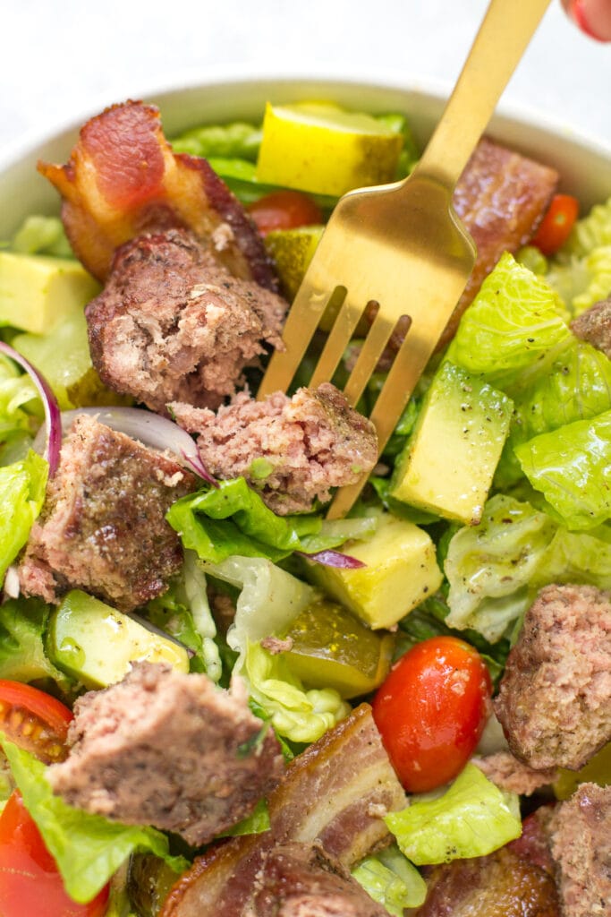 a burger salad in a white bowl with a golden fork taking a bite out of it.