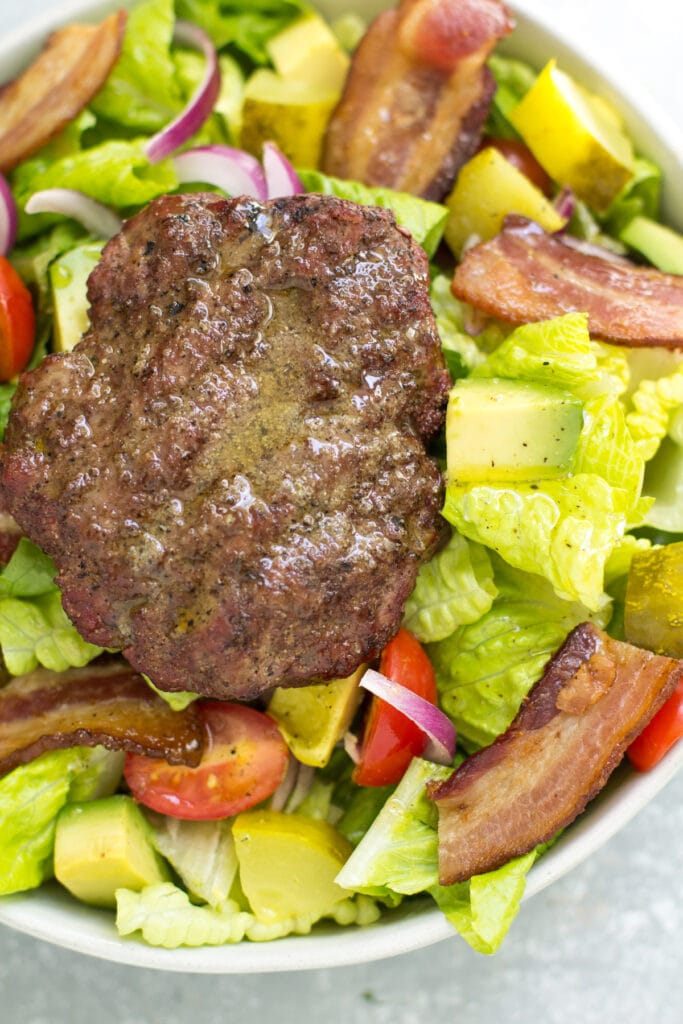 A white bowl filled with keto burger salad. A bed of romaine with bacon, tomatoes, pickles and red onion