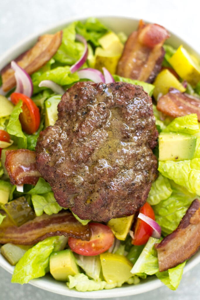 A low carb, whole30 burger salad in a white bowl with bacon, avocado, red onion, cherry tomatoes and a juicy patty on top