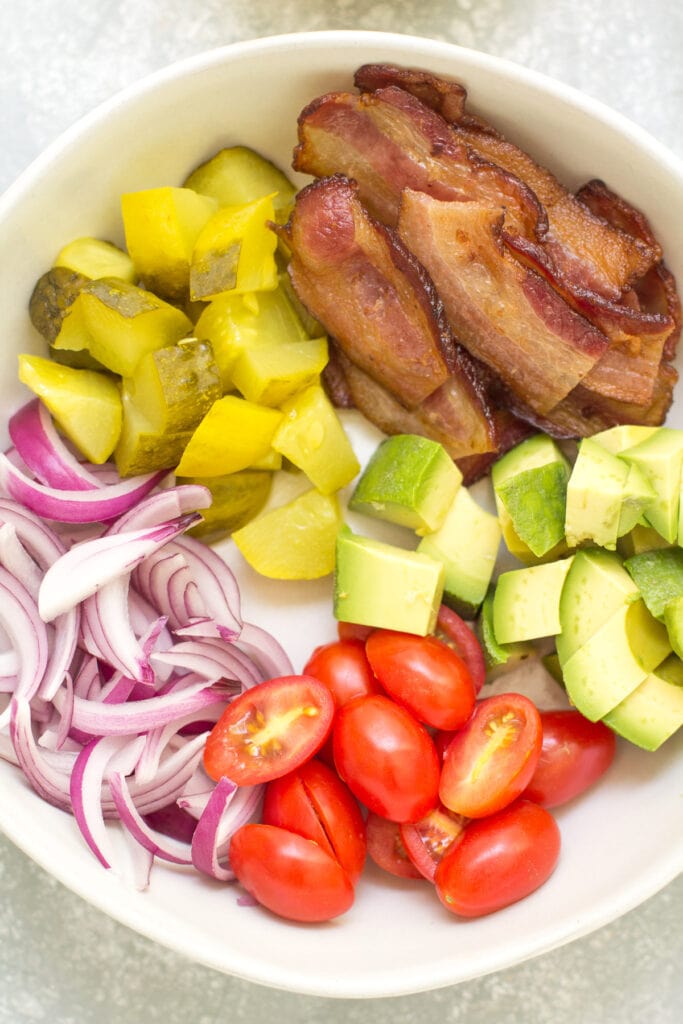 Ingredients for a burger salad in a white bowl. All ingredients are low carb, whole30, paleo and keto: Tomatoes, red onion, avocado, chopped dill pickles, crispy bacon