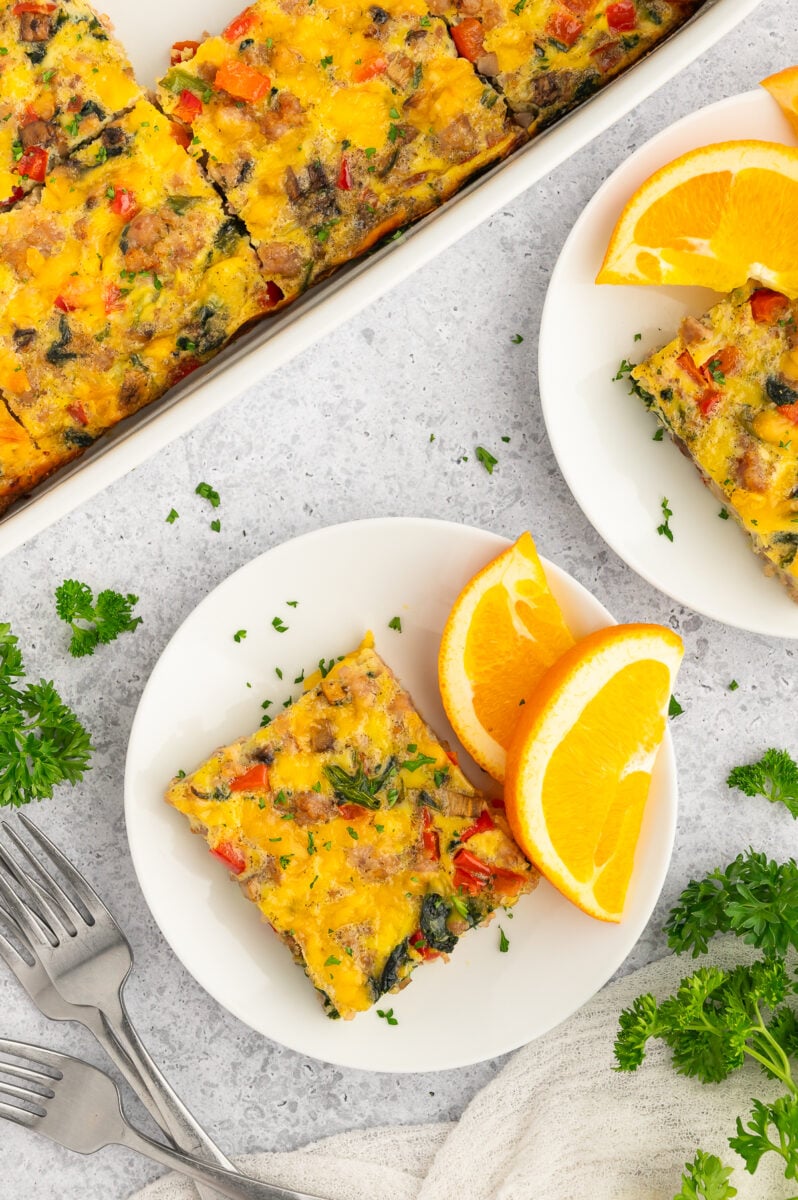 A serving of sausage egg bake on a white plate with orange slices and the casserole dish on the side.
