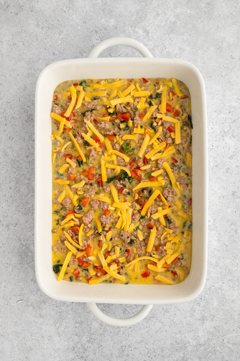 Uncooked sausage egg bake in a white casserole dish topped with cheddar cheese.