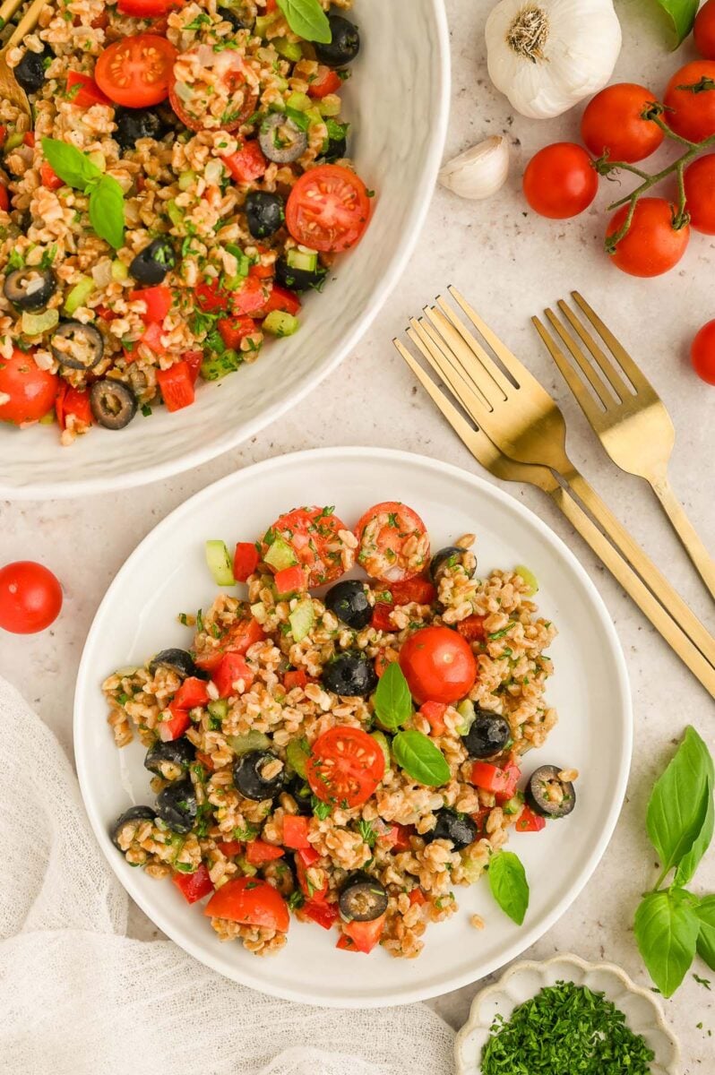 Easy Farro Salad on a white plate with a large bowl of the salad and gold forks on the side. 