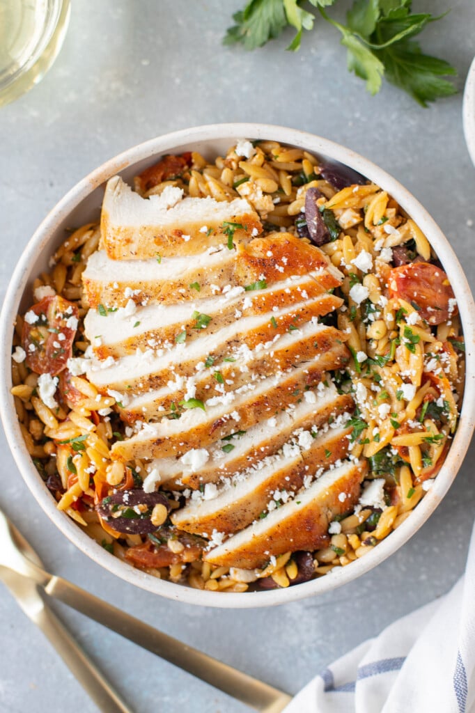 A piece of sliced mediterranean chicken on top of mediterranean orzo salad on a grey background