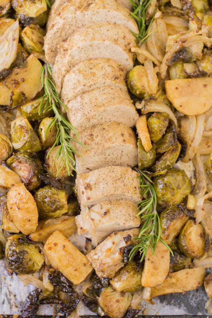 A top down image of Apple pork tenderloin sliced on a baking sheet with brussels sprouts, onions and apples all around.