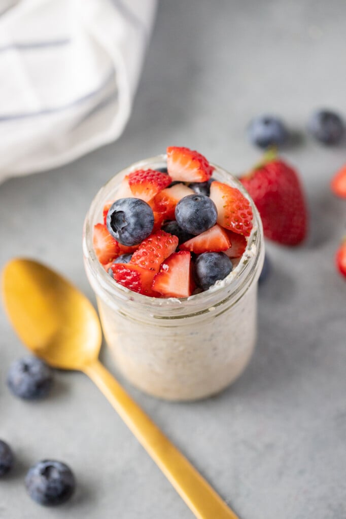 berry overnight oats in a mason jar with berries and a gold spoon on the side on a grey background