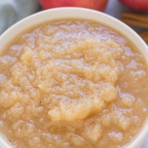 Instant pot applesauce in a white bowl on a blue background with apples and spices in the background.