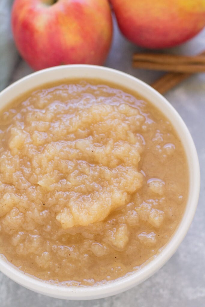 homemade Applesauce in a white bowl on a grey background. Apples and cinnamon sticks are in the background.