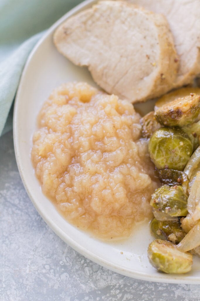 Instant Pot applesauce on a white dish with pork and brussels sprouts. A blue linen is in the background