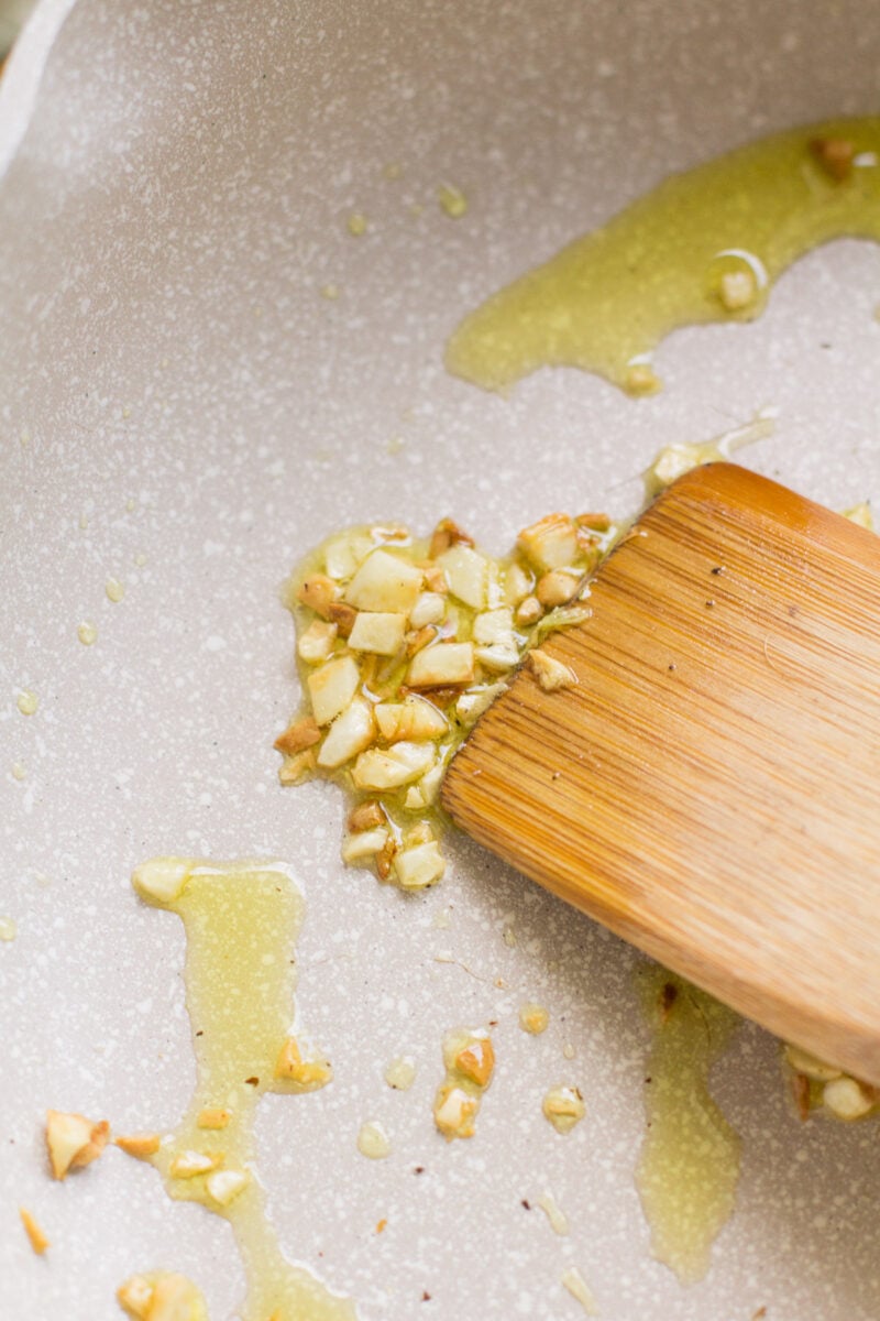Garlic getting sautéed in olive oil in a pan with a wooden spoon.