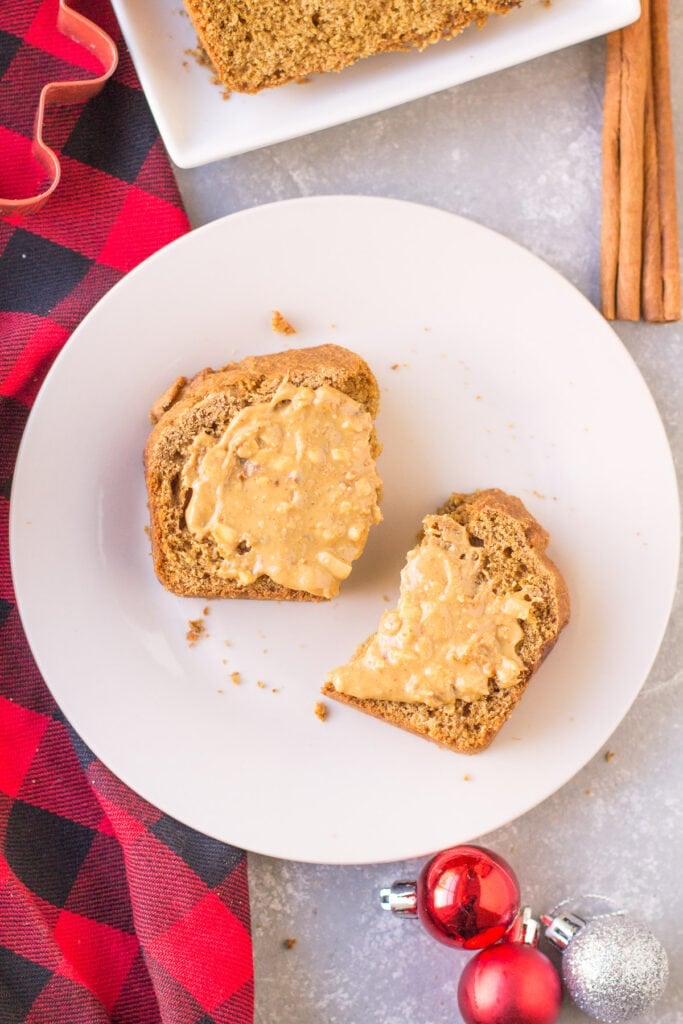 A slice of healthy gingerbread broken in half and covered with peanut butter on a white plate with a red linen and cinnamon sticks off to the side