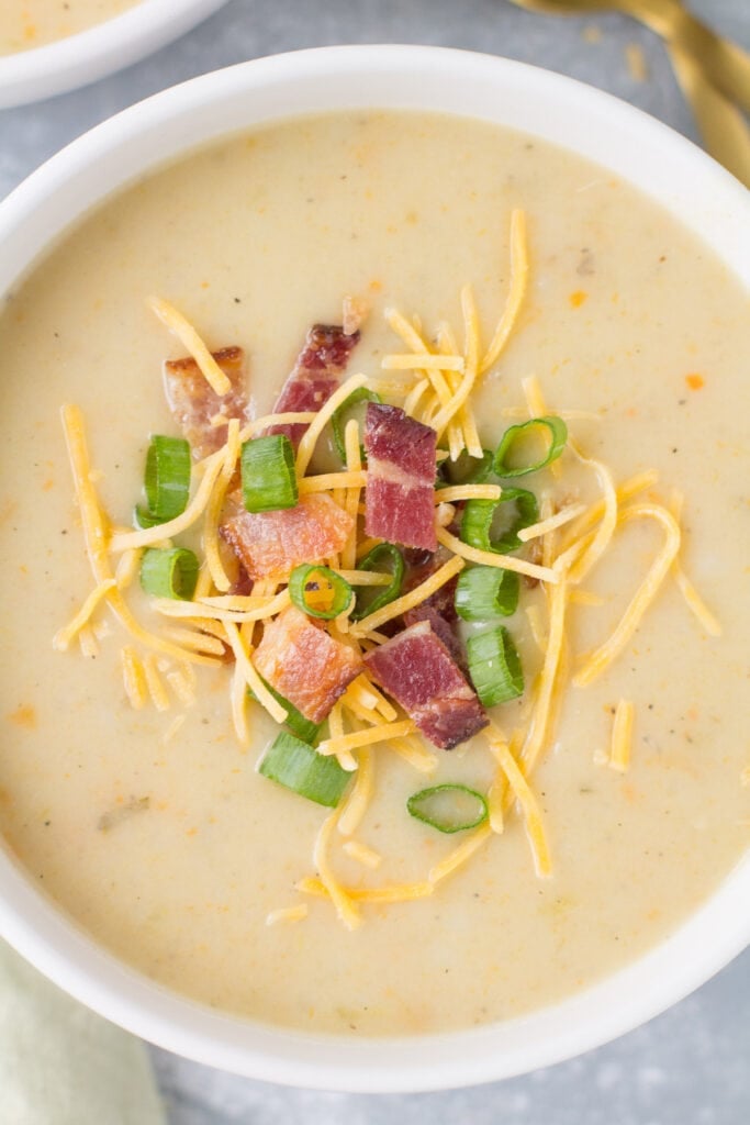 A close up of healthy potato soup on a grey background. The creamy potato soup is topped with bacon, cheddar cheese + chives