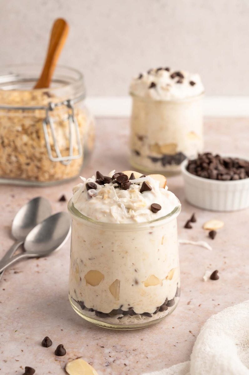 Healthy Almond Joy Overnight Oats in a clear glass jar with almonds and mini chocolate chips scattered around and on top with another jar in the background.