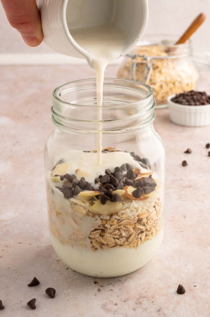 Almond Joy Overnight Oats in a clear jar before being mixed together with milk being poured in.