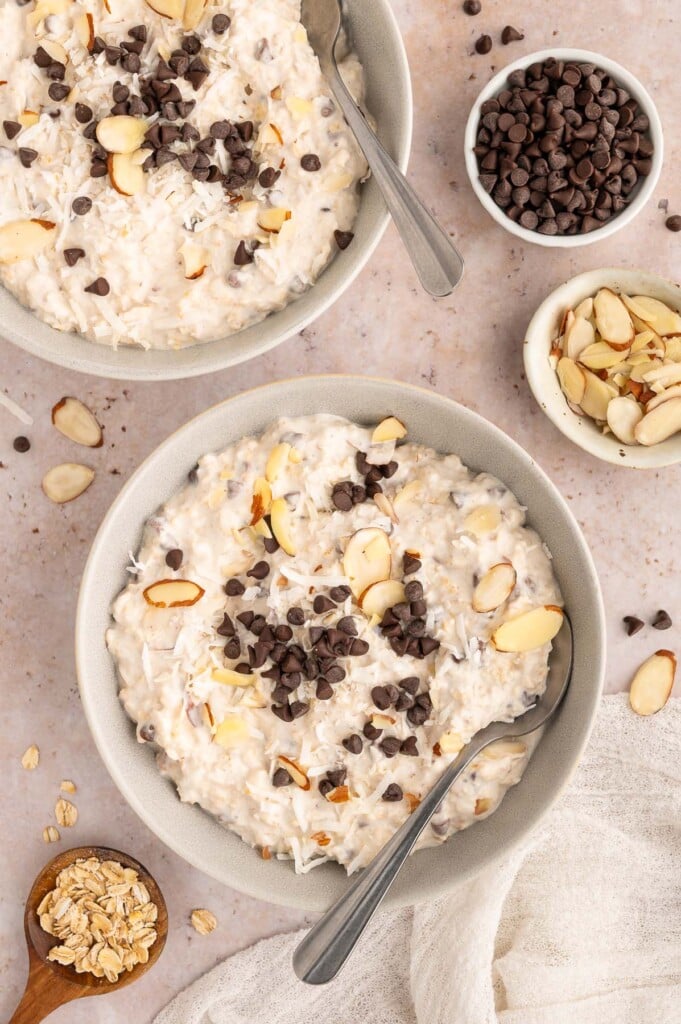 An image of a big bowl of healthy almond joy overnight oats with another bowl, chocolate chips, almonds, and oats scattered around.
