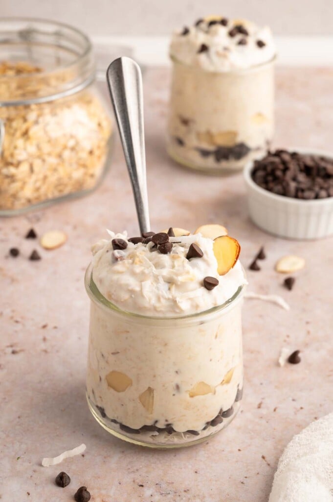 Healthy Almond Joy Overnight Oats in a clear glass jar with a spoon in the jar and almonds and mini chocolate chips scattered around and on top with another jar in the background.