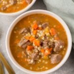 Healthy Beef Barley Soup in white bowls with a green linen and gold spoons off to the side.