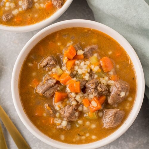 Healthy Beef Barley Soup in white bowls with a green linen and gold spoons off to the side.
