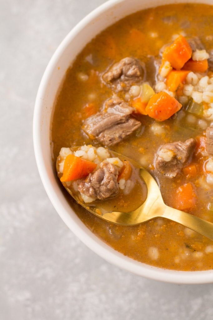 An overhead shot of beef barley soup with a gold spoon taking a bite out of it.