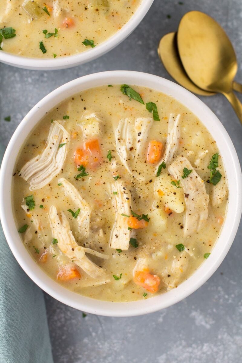 Healthy Chicken Pot Pie Soup in a white bowl on a grey background. Gold spoons are off to the right side