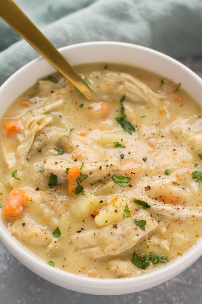 A bowl of chicken pot pie soup with black pepper on top in a grey background with a green linen. A gold spoon is stirring the soup.