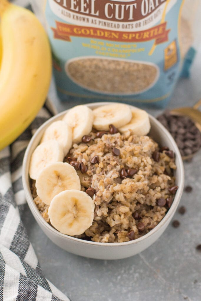 A close up of baked steel cut oatmeal with bananas on a grey background white a bag of oats, bananas and chocolate in the background.