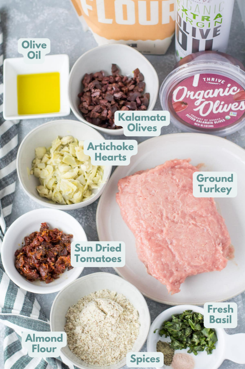 Ingredients for healthy Meatballs laid out in small white bowls With greek flavors and made with turkey: kalamata olives, olive oil, artichoke hearts, sun dried tomatoes, almond flour, ground turkey, dried oregano, fresh parsley, pepper