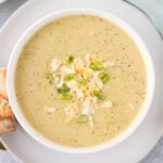 An overhead shot of Healthy Broccoli Soup in a white bowl on a white plate with bread off to the side.