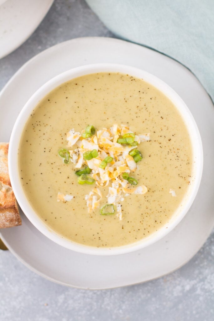 An overhead shot of Healthy Broccoli Soup in a white bowl on a white plate with bread off to the side.
