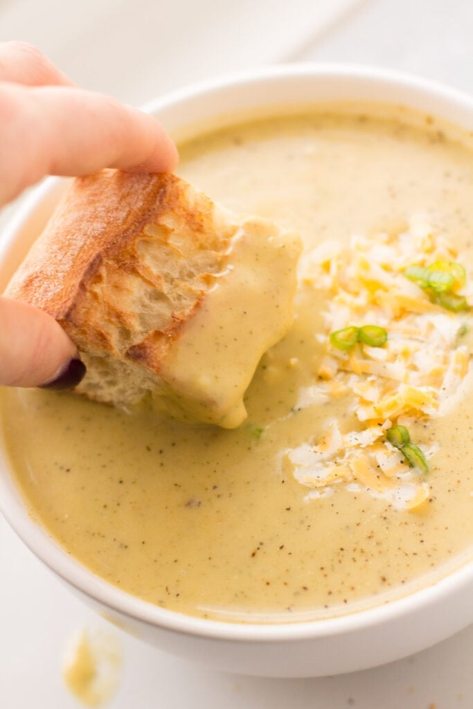 A hand dunking a piece of bread into a bowl of healthy broccoli soup
